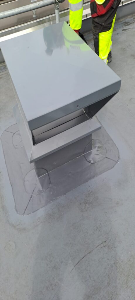 Metal vent on grey rooftop with worker's leg visible.
